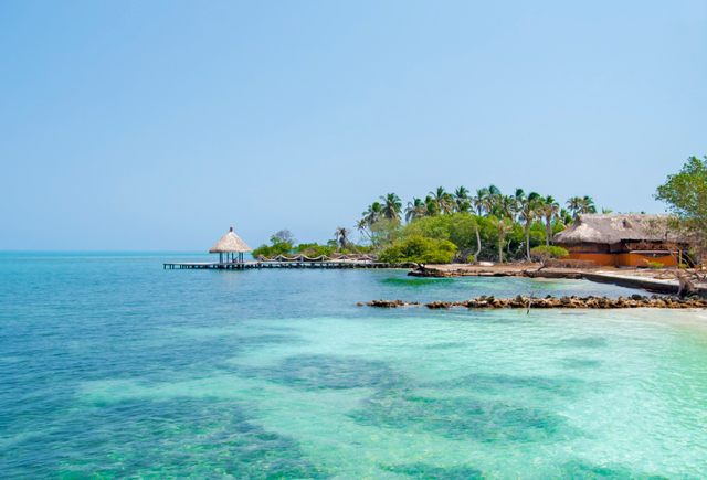Plage paradisiaque en Colombie avec eau turquoise, palmiers, ponton en bois et cabanes tropicales.